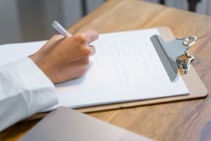 A close-up of a person's hand writing on a clipboard on a wooden table, displaying focus and concentration.